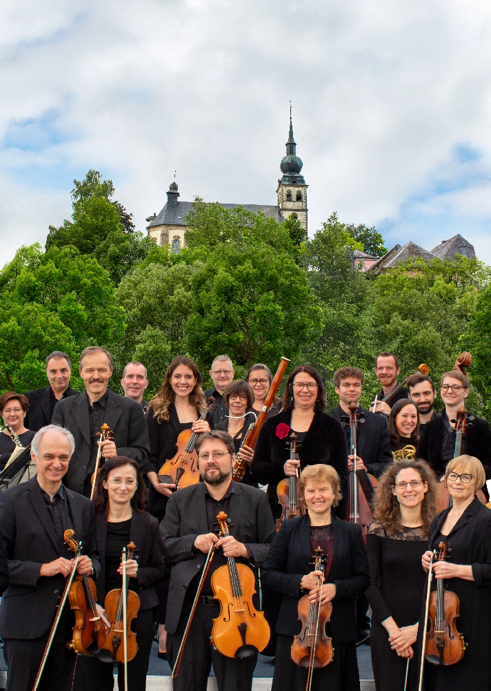 Smiling musicians from the Estro Armonico orchestra stand with their instruments under a cloudy sky with a church in the background.